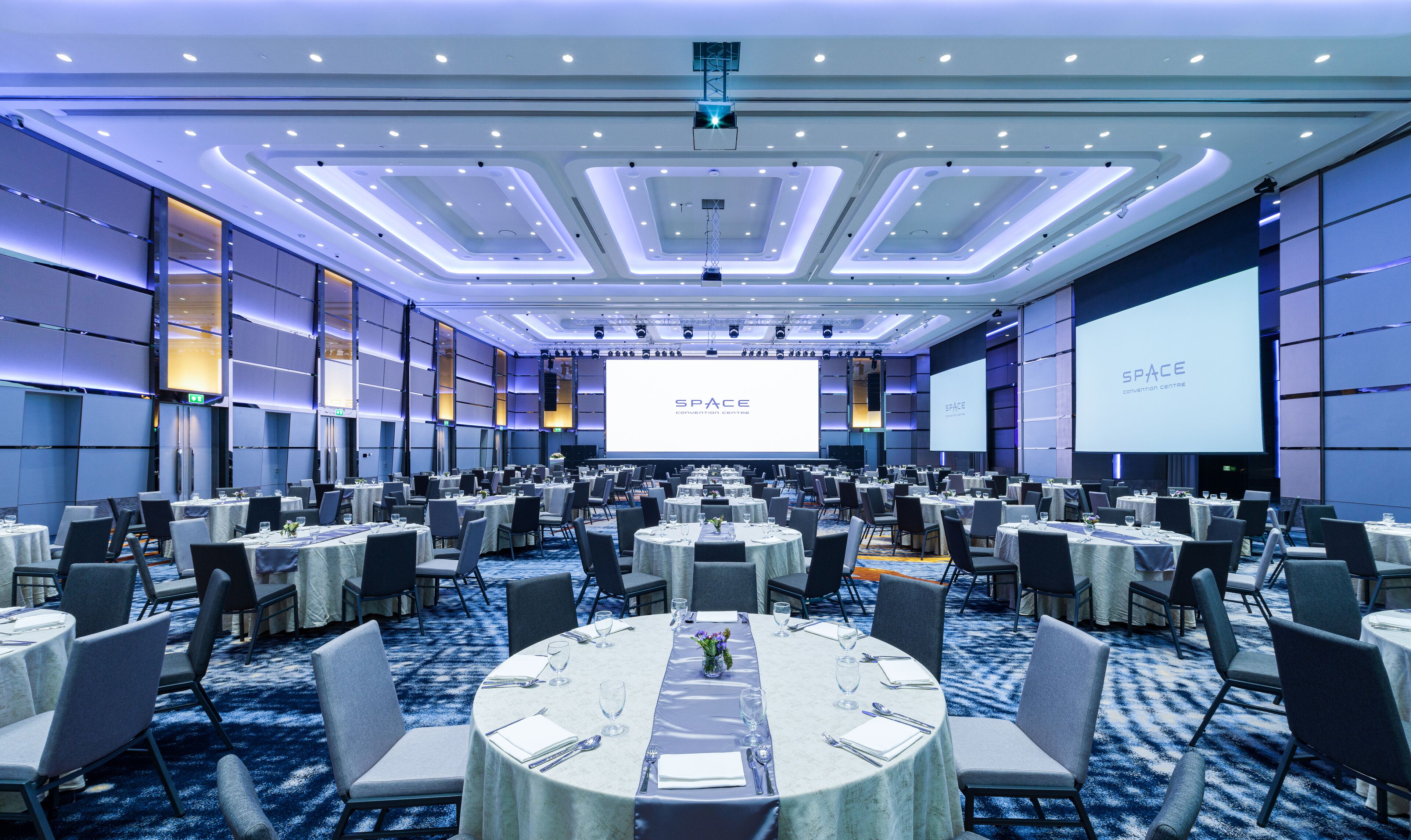 Un salón de conferencias amplio con varias mesas y sillas, una pantalla grande que muestra 'SPACE Convention Centre' y un diseño de techo moderno.