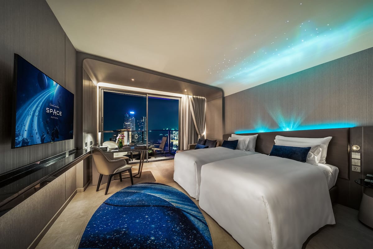 A modern hotel room with a large TV displaying a space-themed movie, a balcony with a city view, and a starry night sky projection on the ceiling.