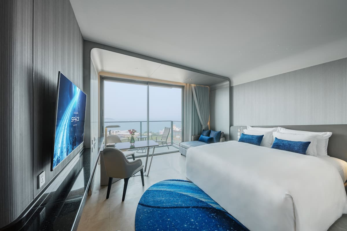 A modern hotel room with a large bed, a TV, a balcony with a view, and a desk with a chair.