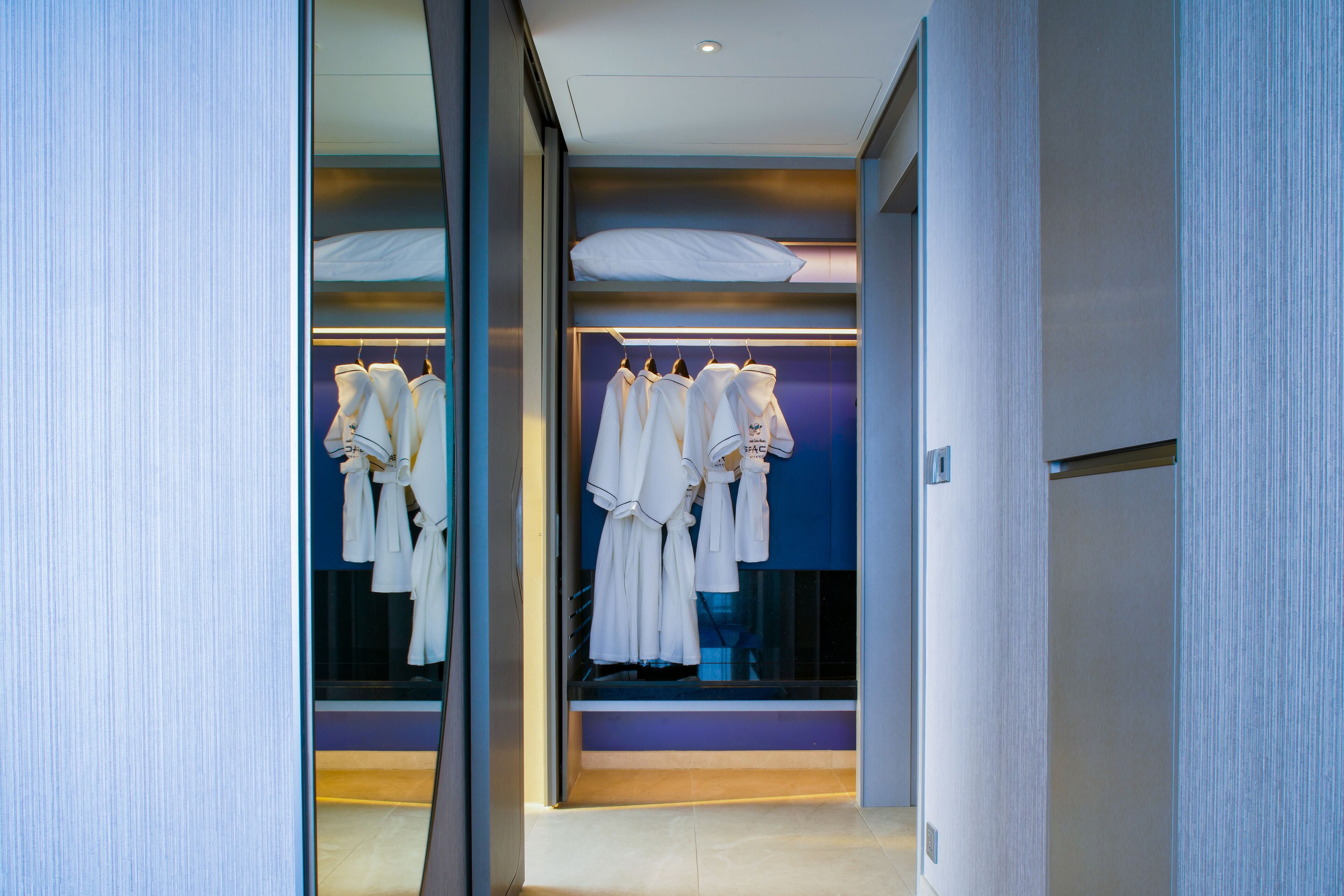 Habitación de hotel moderna con vestidor que contiene batas y toallas blancas ordenadas.