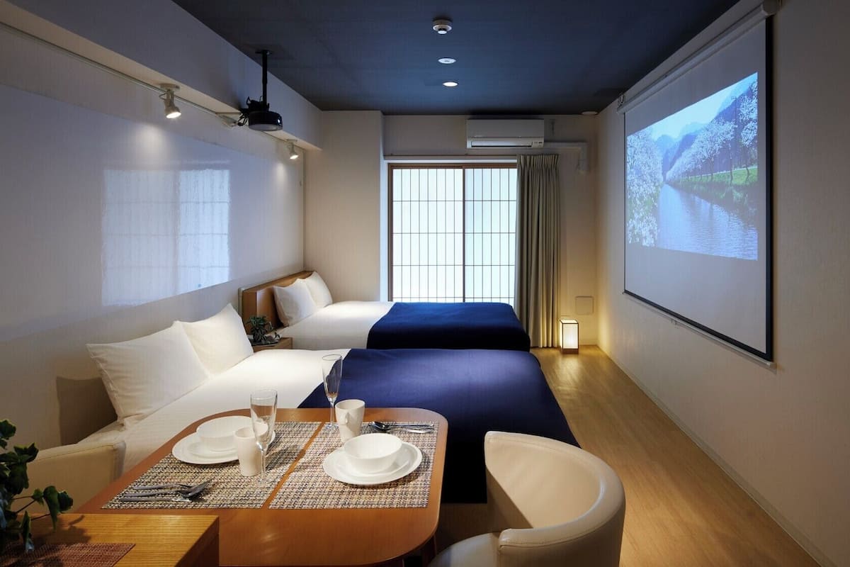 Une chambre d’hôtel comprenant un lit, un bureau avec une chaise, une table à manger dressée pour deux personnes et un grand écran diffusant un paysage pittoresque.