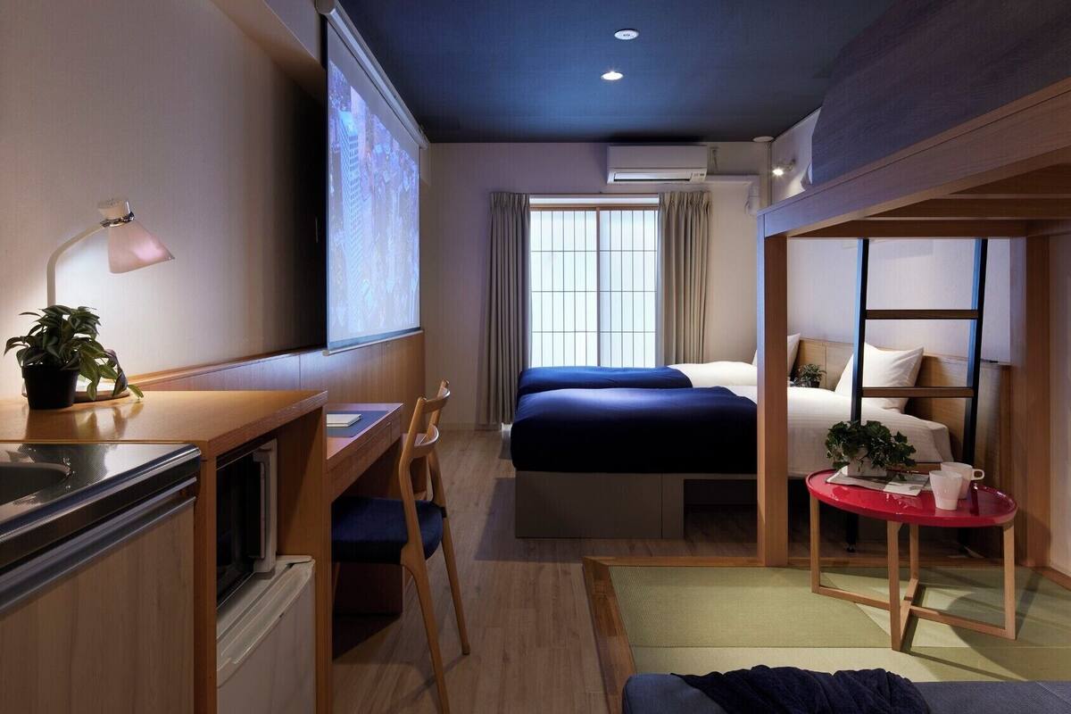 Une chambre d’hôtel compacte comprenant un lit, un bureau avec une chaise, une petite table avec une plante et un écran de projection.