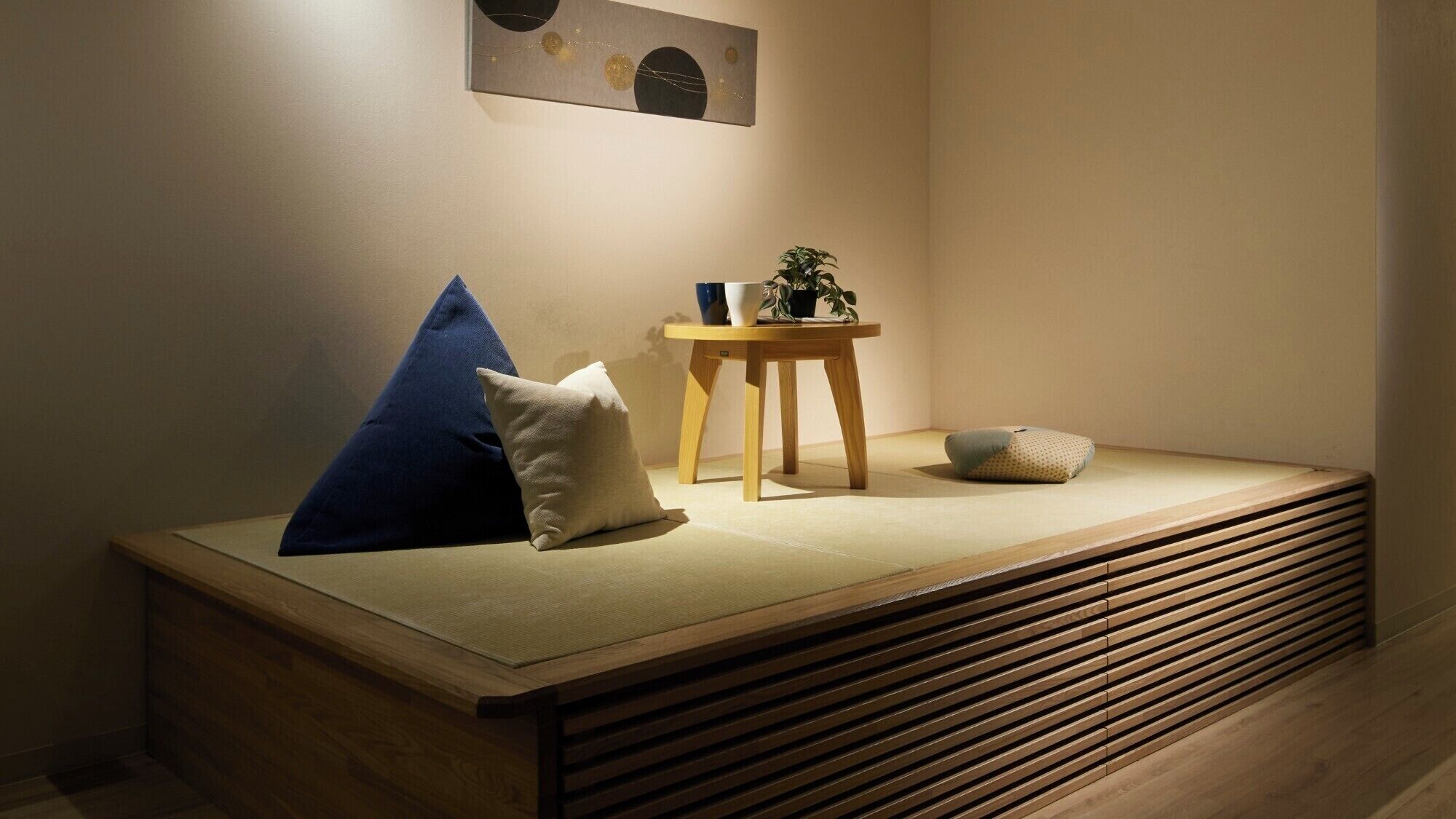 A minimalist room with a low wooden bed, a small wooden table with a plant, and a wall-mounted abstract art piece.