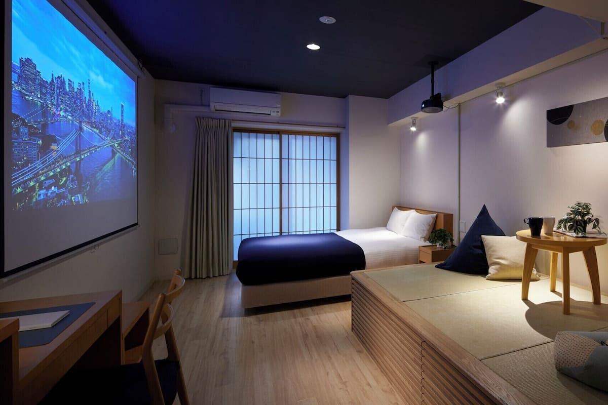 Une chambre d’hôtel moderne équipée d’un lit, d’un bureau, d’un écran de projection diffusant une vue sur la ville, et d’une petite table agrémentée d’une plante.
