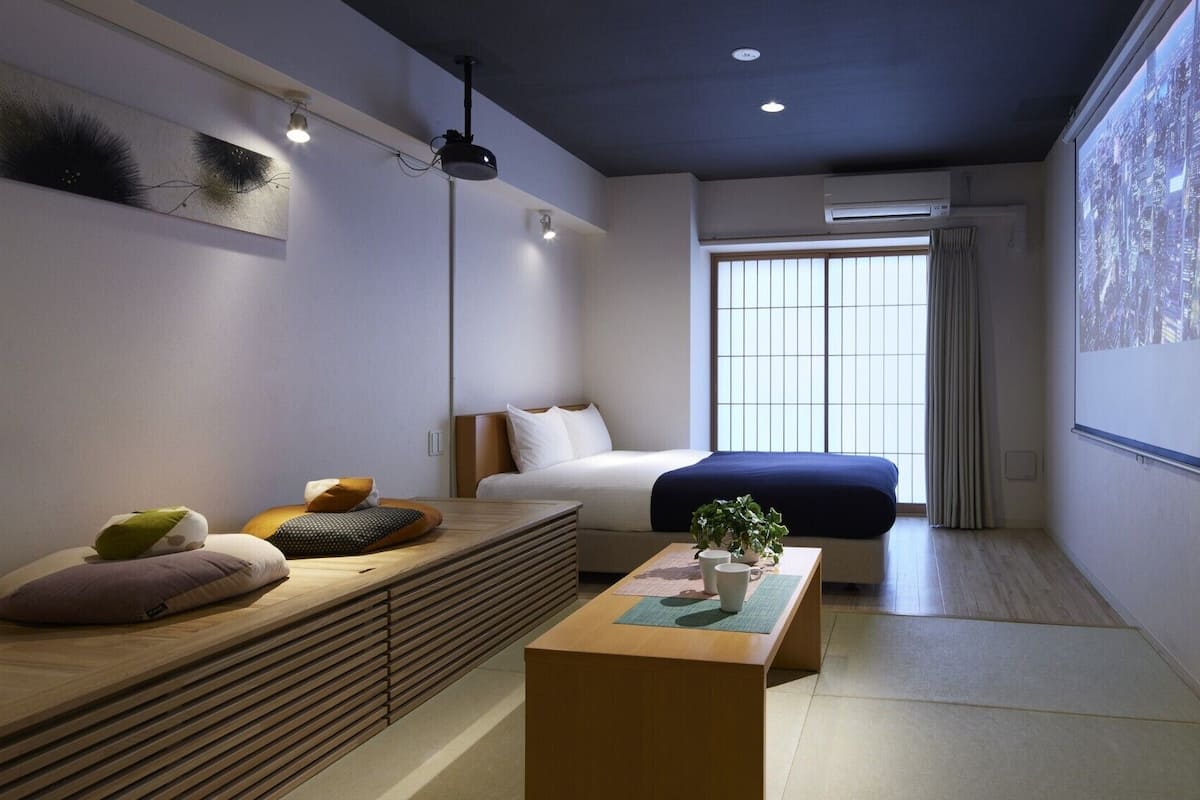 Une chambre d’hôtel moderne, dotée d’un lit, d’un coin salon avec des coussins, d’une table basse et d’une grande fenêtre offrant une vue sur le paysage urbain.