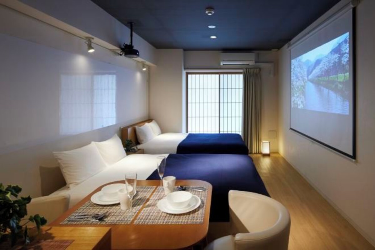 A hotel room with a bed, a desk with a chair, a dining table set, and a large screen displaying a scenic view.