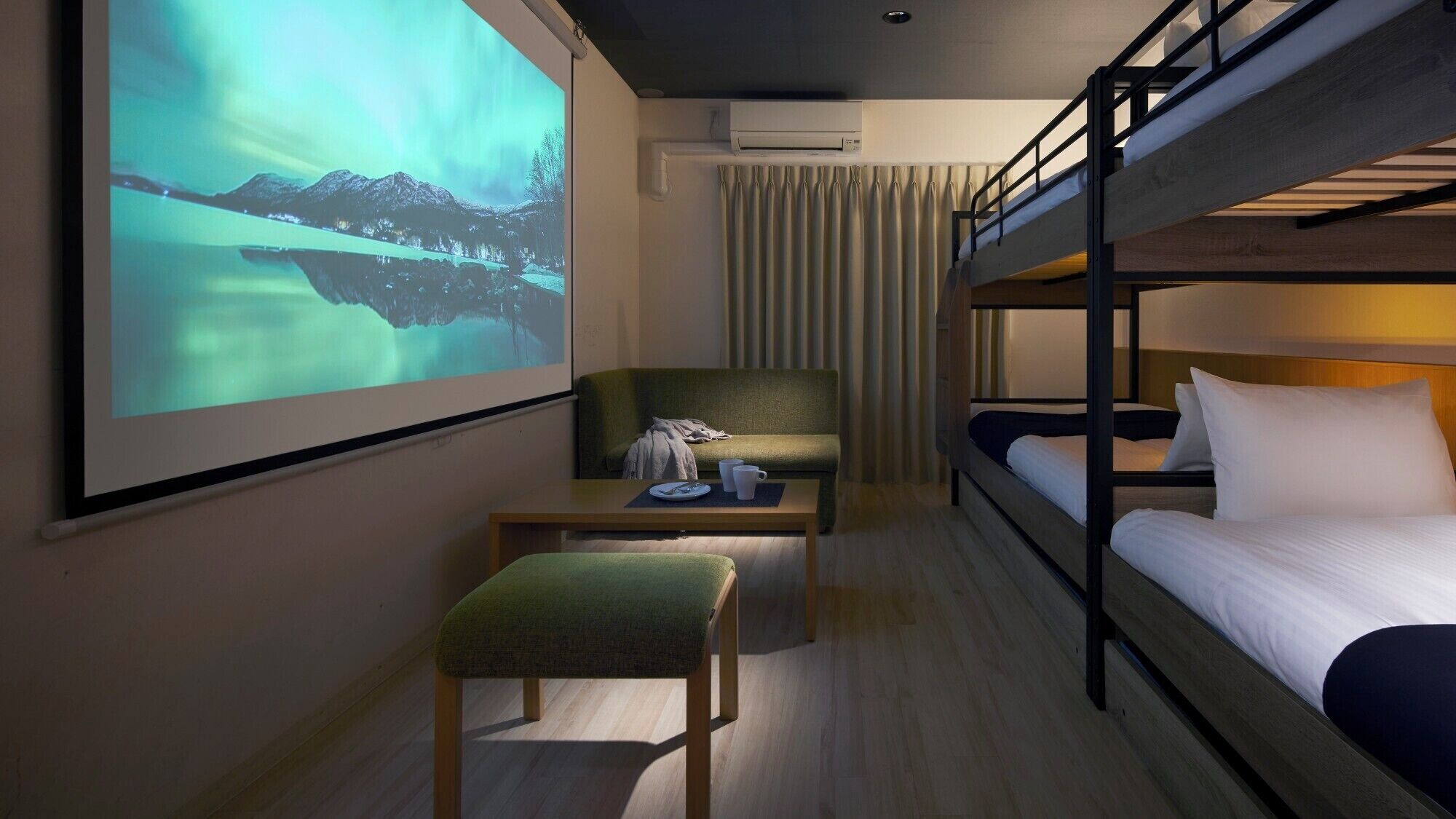 A hotel room with a bunk bed, a desk with a chair, a projector screen displaying a landscape, and an air conditioning unit.