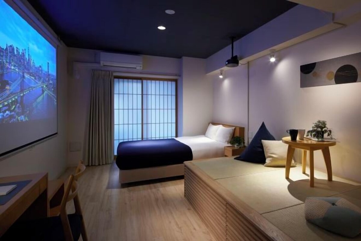 A modern hotel room with a bed, a sofa, a desk, and a projector screen displaying a cityscape.