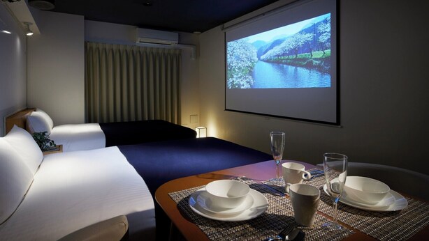 Habitación de hotel con cama, escritorio con mesa de comedor y pantalla proyectando un paisaje.