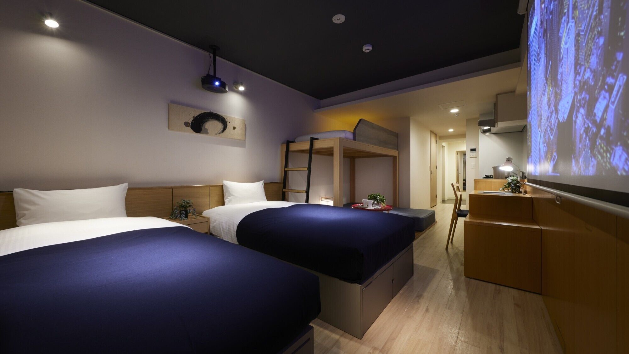 A hotel room with two beds, a bunk bed, a desk, a chair, a small table, and a projector screen displaying a cityscape.