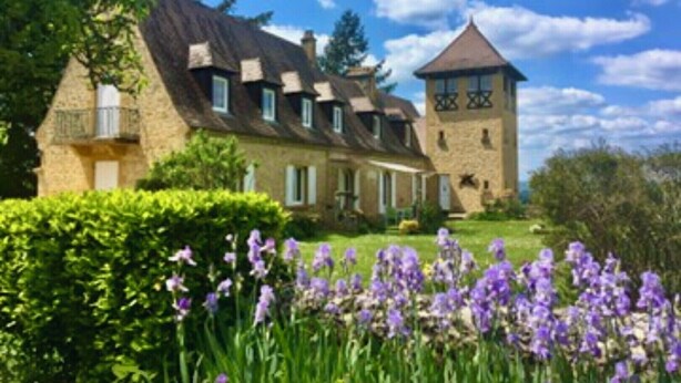 A large stone building with multiple chimneys, a tower, and a well-maintained garden with purple flowers. - Magnificent Villa on 50 ha overlooking the medieval town of Domme (Domme)