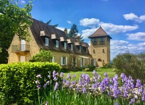 Un grande edificio in pietra con diverse canne fumarie, una torre e un giardino curato con fiori viola.