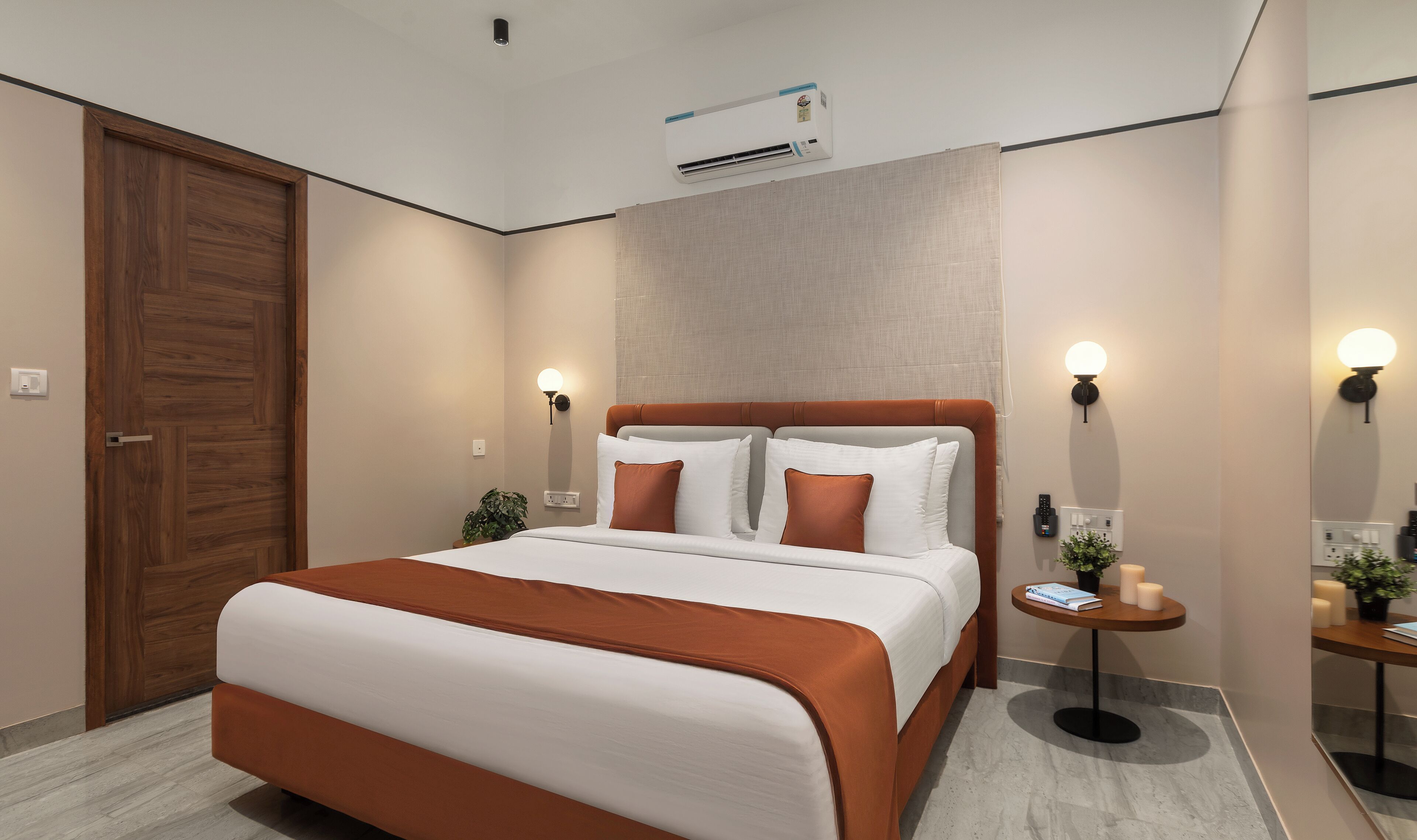 Une chambre moderne avec un grand lit, une porte en bois, un climatiseur et une table de chevet agrémentée d’une plante et de bougies.