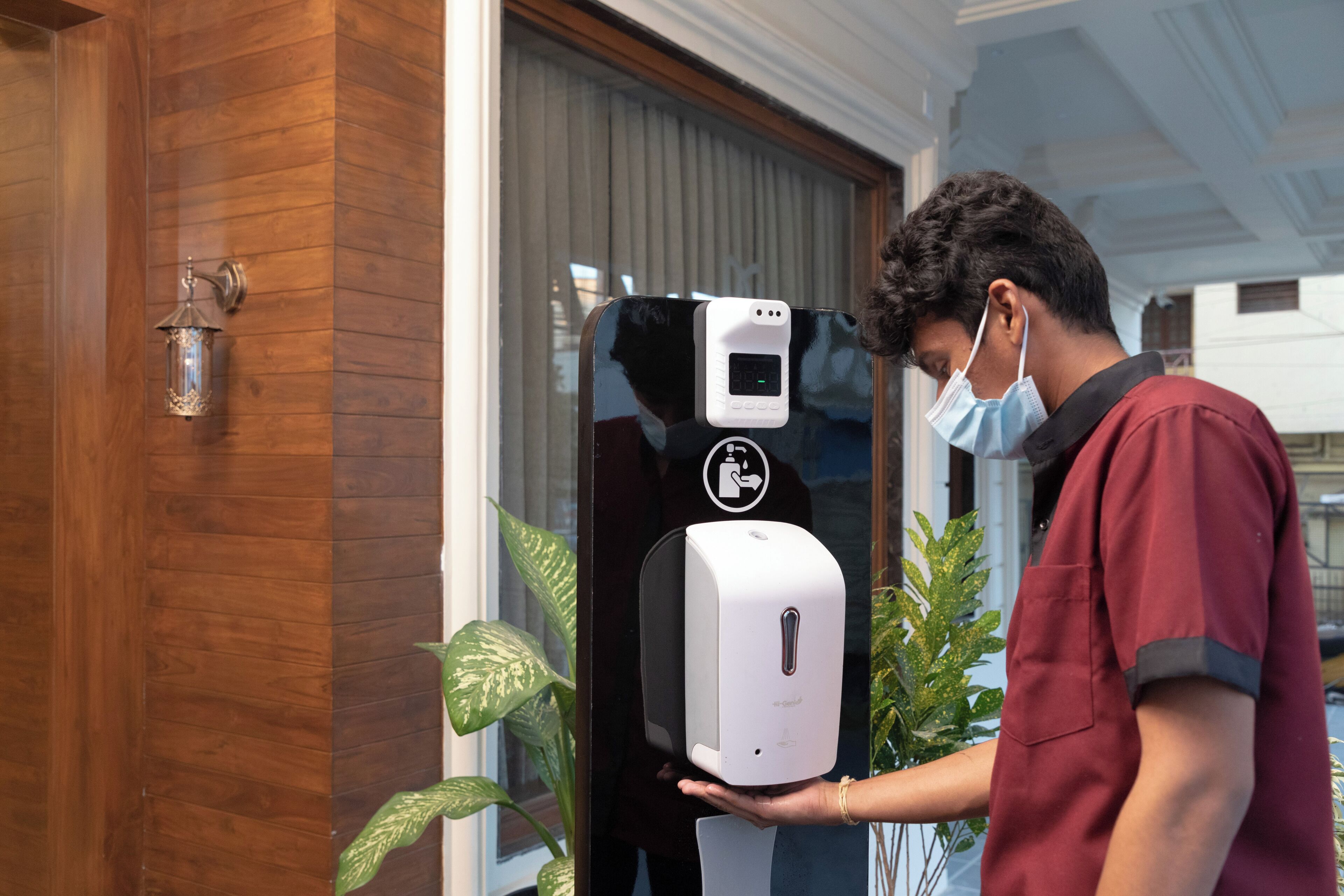A person using a hand sanitizer dispenser mounted on a wooden wall near a window.