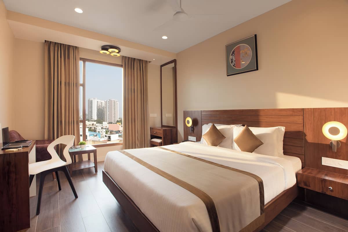 A hotel room with a large bed, a desk with a chair, a small table with a plant, and a view of the city through large windows.