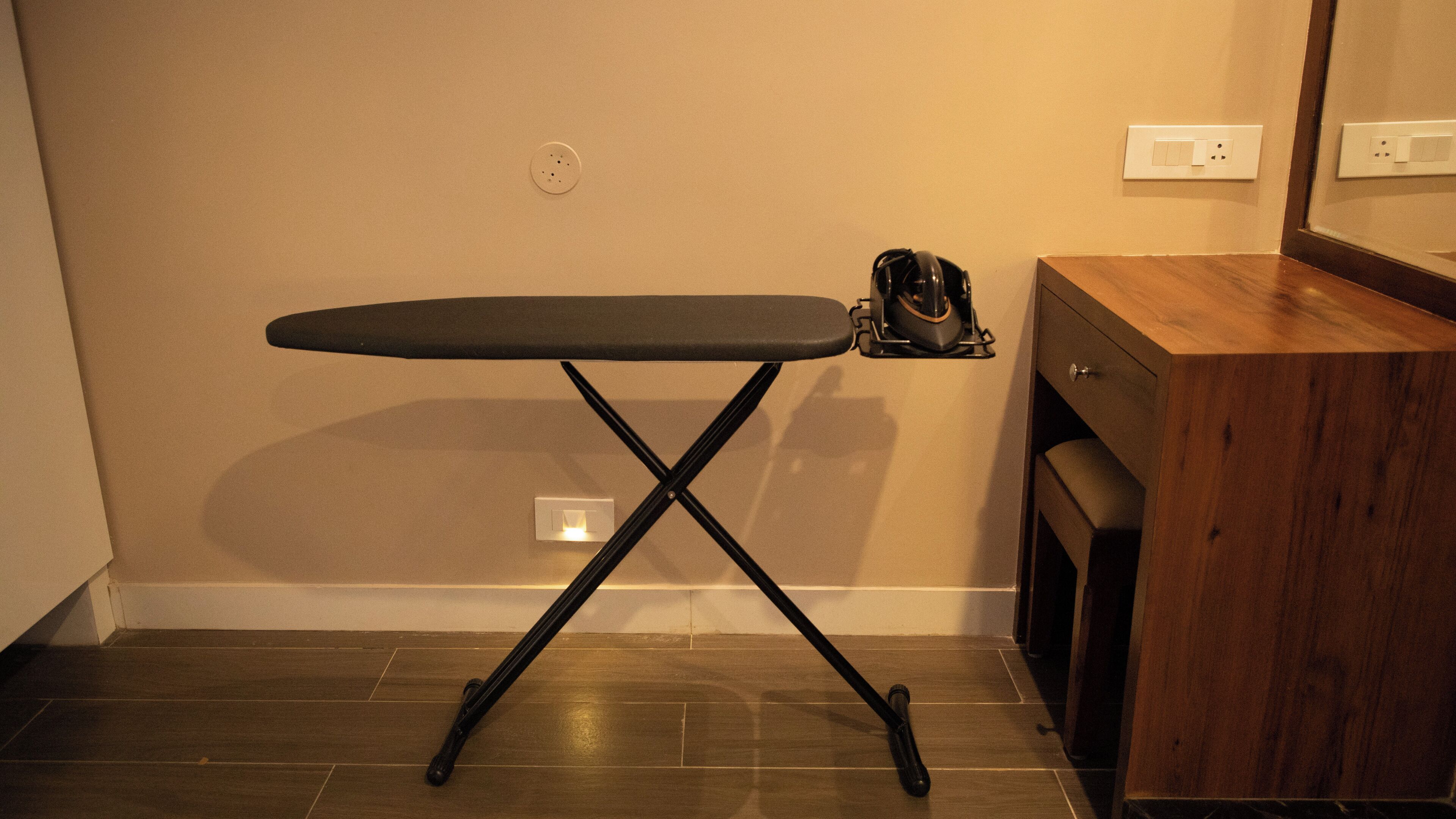 A compact ironing board with a stand, positioned against a wall with a wooden cabinet to the right.