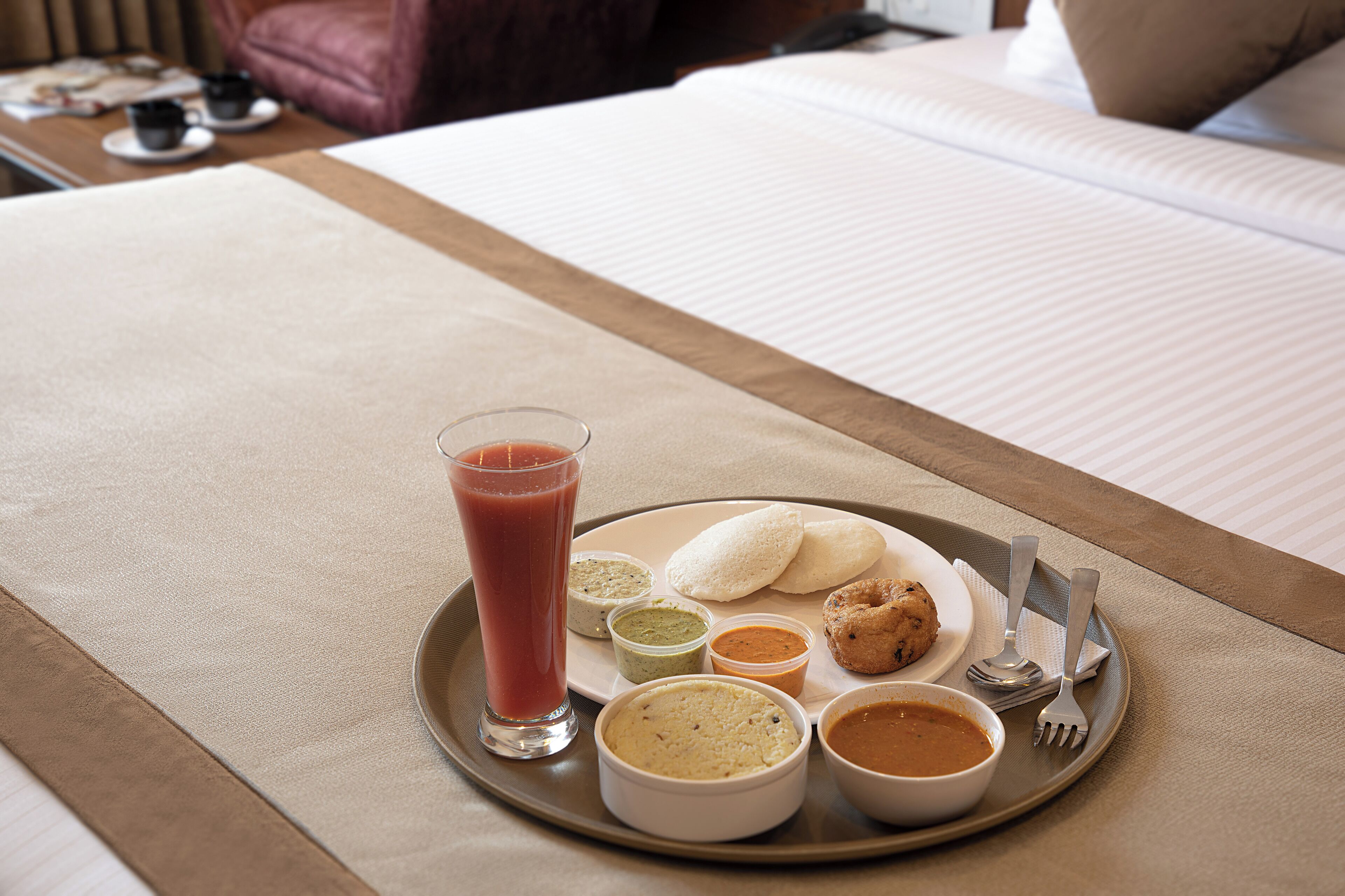 A tray with a glass of smoothie, two idlis, two dosas, and three small bowls of chutney and sambar on a bed.