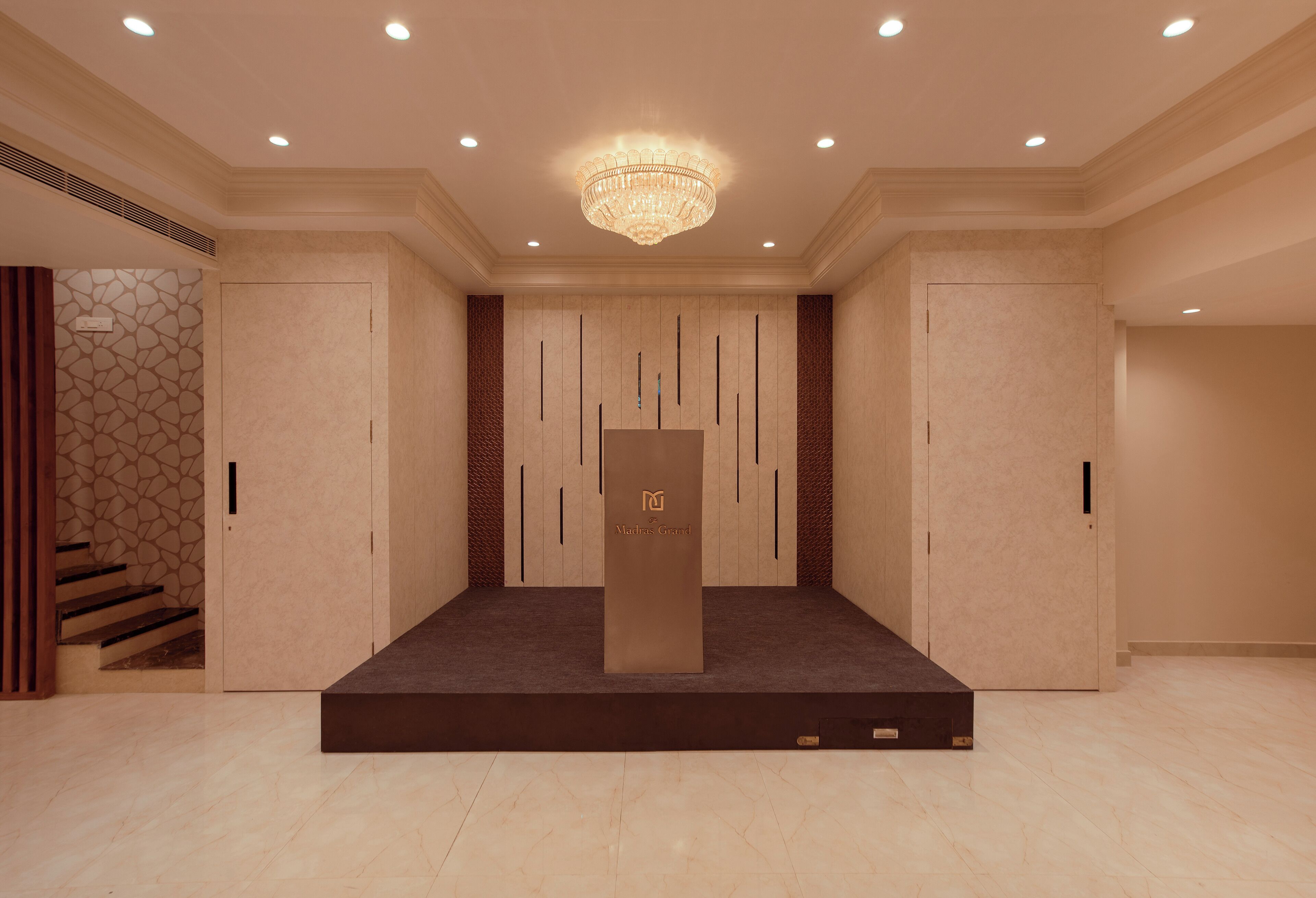 A hotel lobby with a central podium, a chandelier, and a staircase to the left.