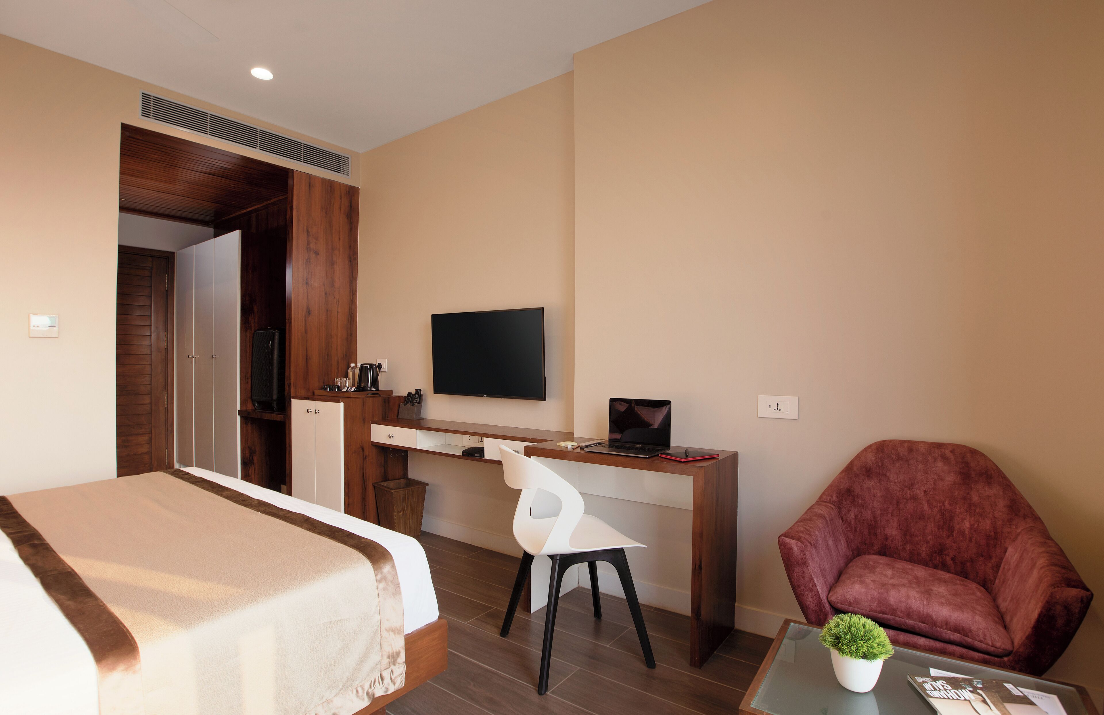 A hotel room with a bed, a desk with a computer, a TV, a red armchair, and a small table with a plant.