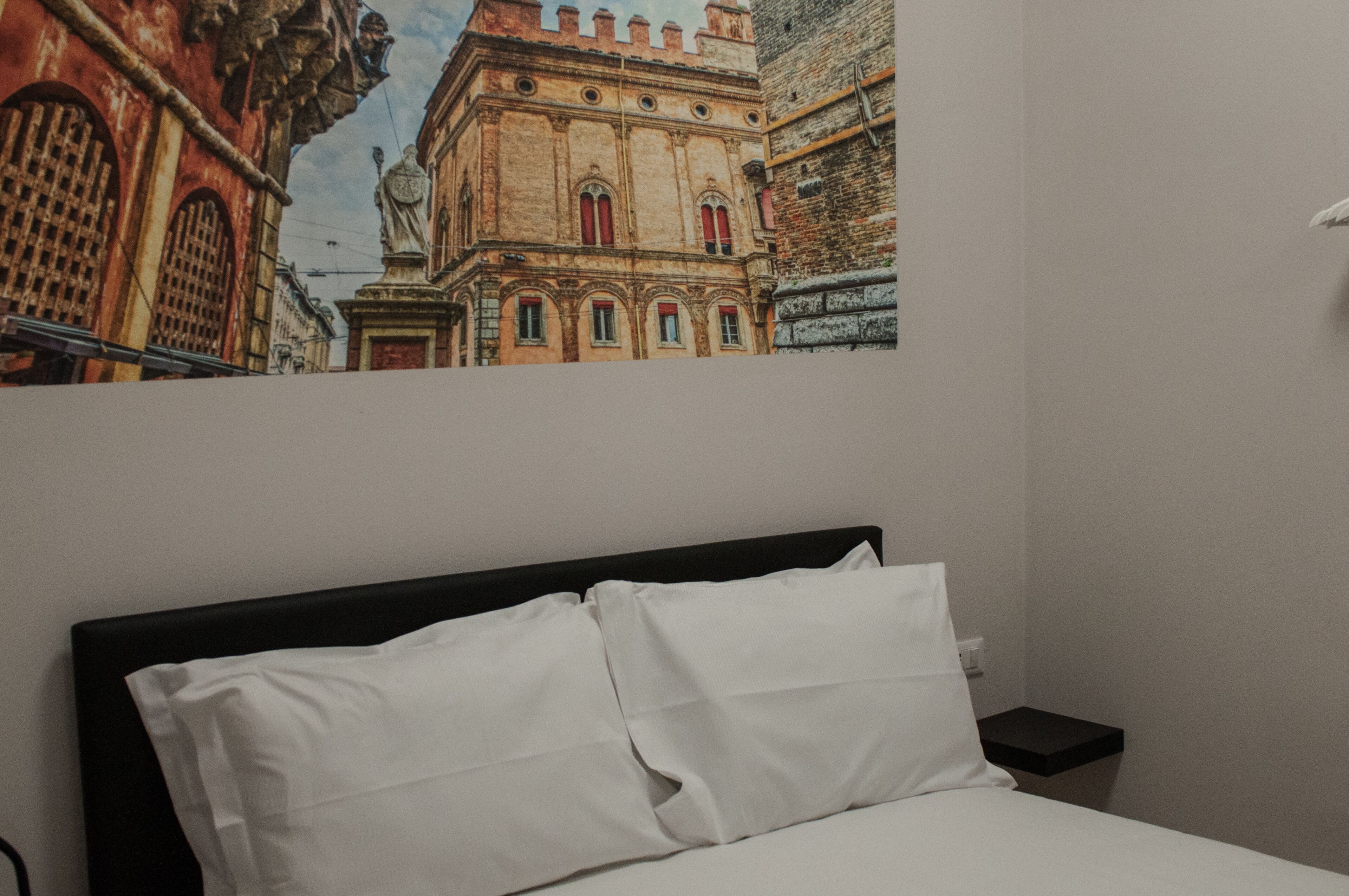 Habitación de hotel con cama, almohadas blancas, mesita de noche y un cuadro colgado en la pared que muestra un edificio.