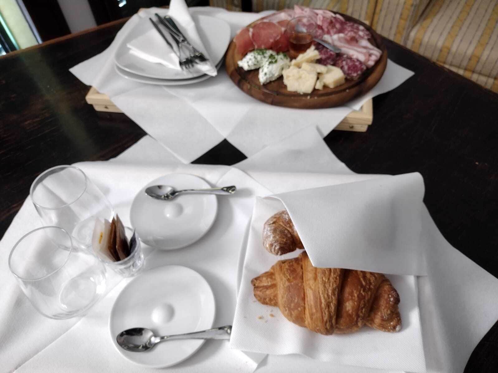 A breakfast setting with a croissant, cheese, and cured meats on a wooden board, accompanied by plates, cutlery, and glasses on a table.