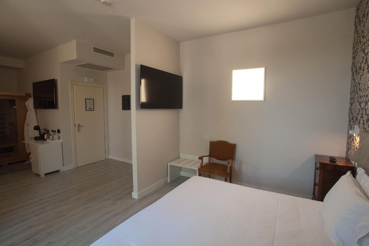 Ein Hotelzimmer mit einem Bett, einem Sessel, einem Schreibtisch, einem an der Wand befestigten Fernseher und einem Fenster mit Tageslicht.