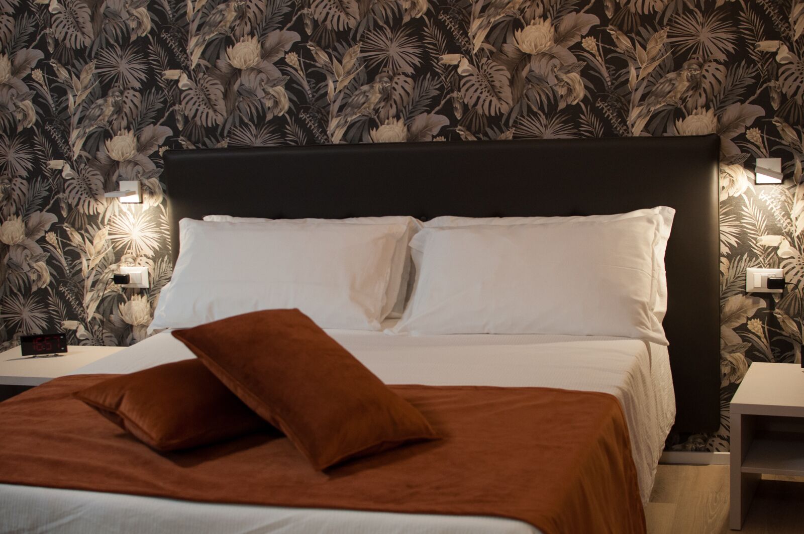 A neatly made bed with white linens and two brown pillows, a black headboard, and a wall with a floral pattern.