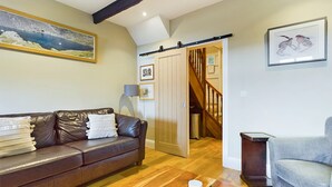 A living room with a brown leather sofa, a grey armchair, wooden flooring, a sliding door, and a staircase. - Stoneswood Cottage, Delph, Saddleworth (Delph)