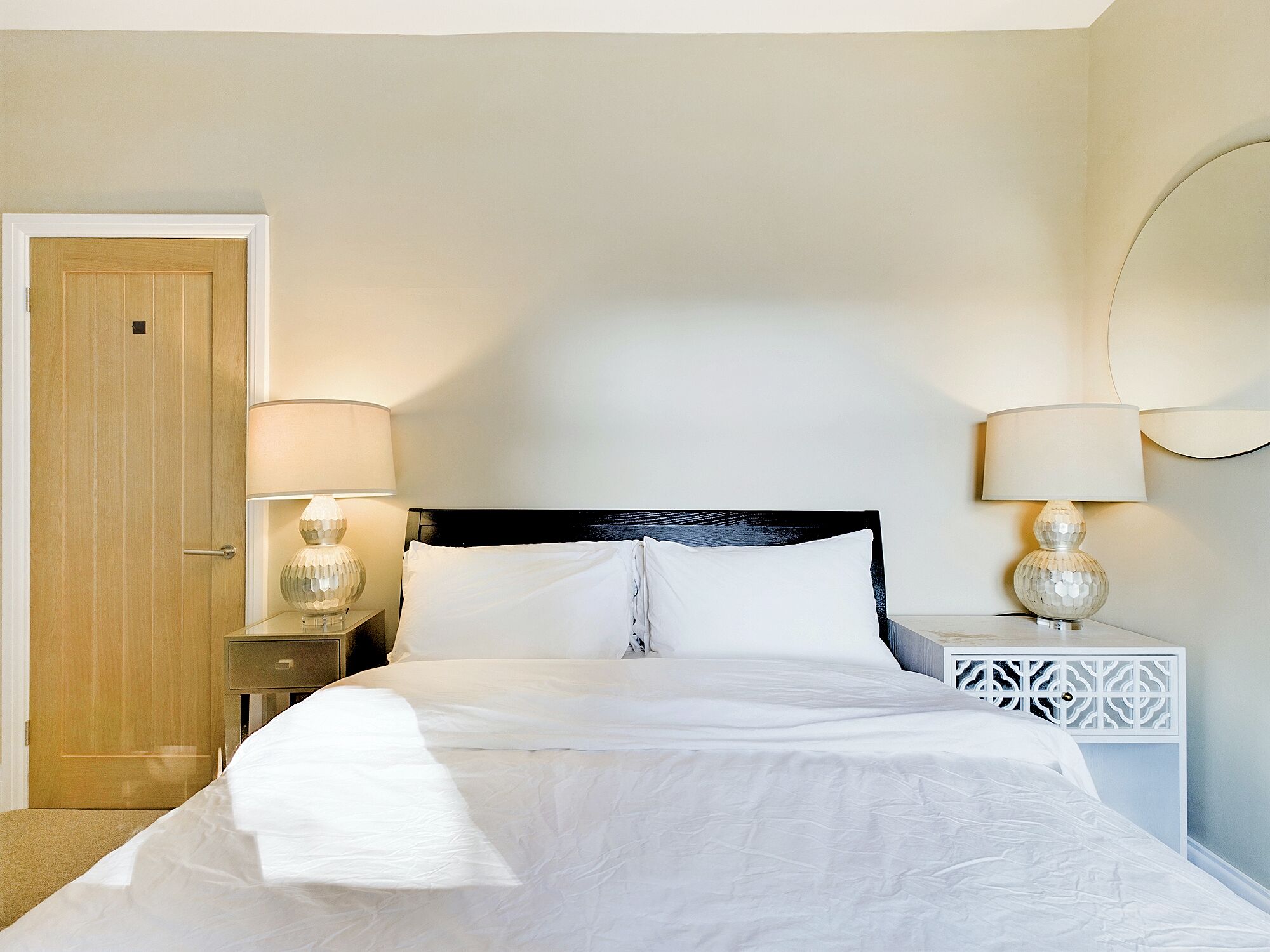 Una cama bien tendida con sábanas blancas, dos lámparas de noche y una puerta de madera.
