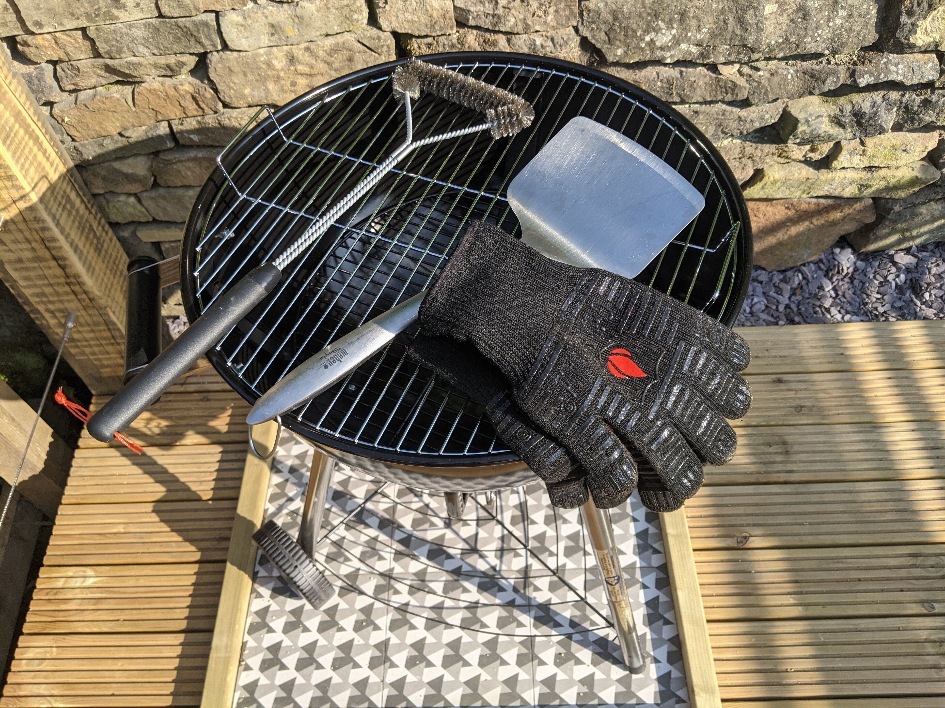 Ein Grill mit einem Metallspatel und einer Bürste, ein Paar Handschuhe und eine Mauer im Hintergrund.