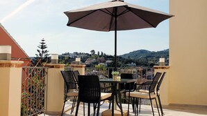 An outdoor patio with a table and chairs, a large umbrella, and a view of a hillside town. - Close to the beach, spacious two-bedroom apartment with breakfast (Corfu)