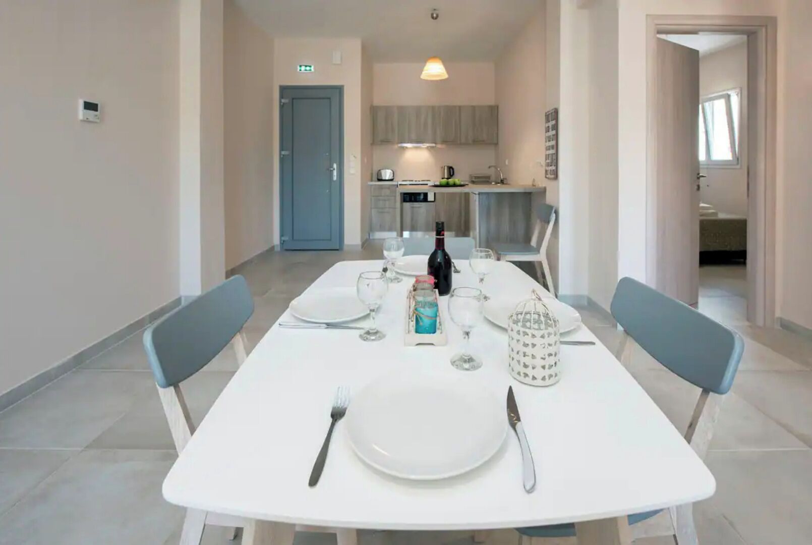 A dining area with a white table set for four, including plates, cutlery, and glasses. A bottle and a decorative item are on the table. The room has a light-colored floor and walls, and there is a door leading to a kitchen area.