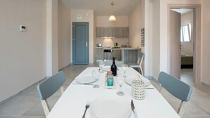 A dining area with a white table set for four, including plates, cutlery, and glasses. A bottle and a decorative item are on the table. The room has a light-colored floor and walls, and there is a door leading to a kitchen area. - Close to the beach, spacious two-bedroom apartment with breakfast (Corfu)