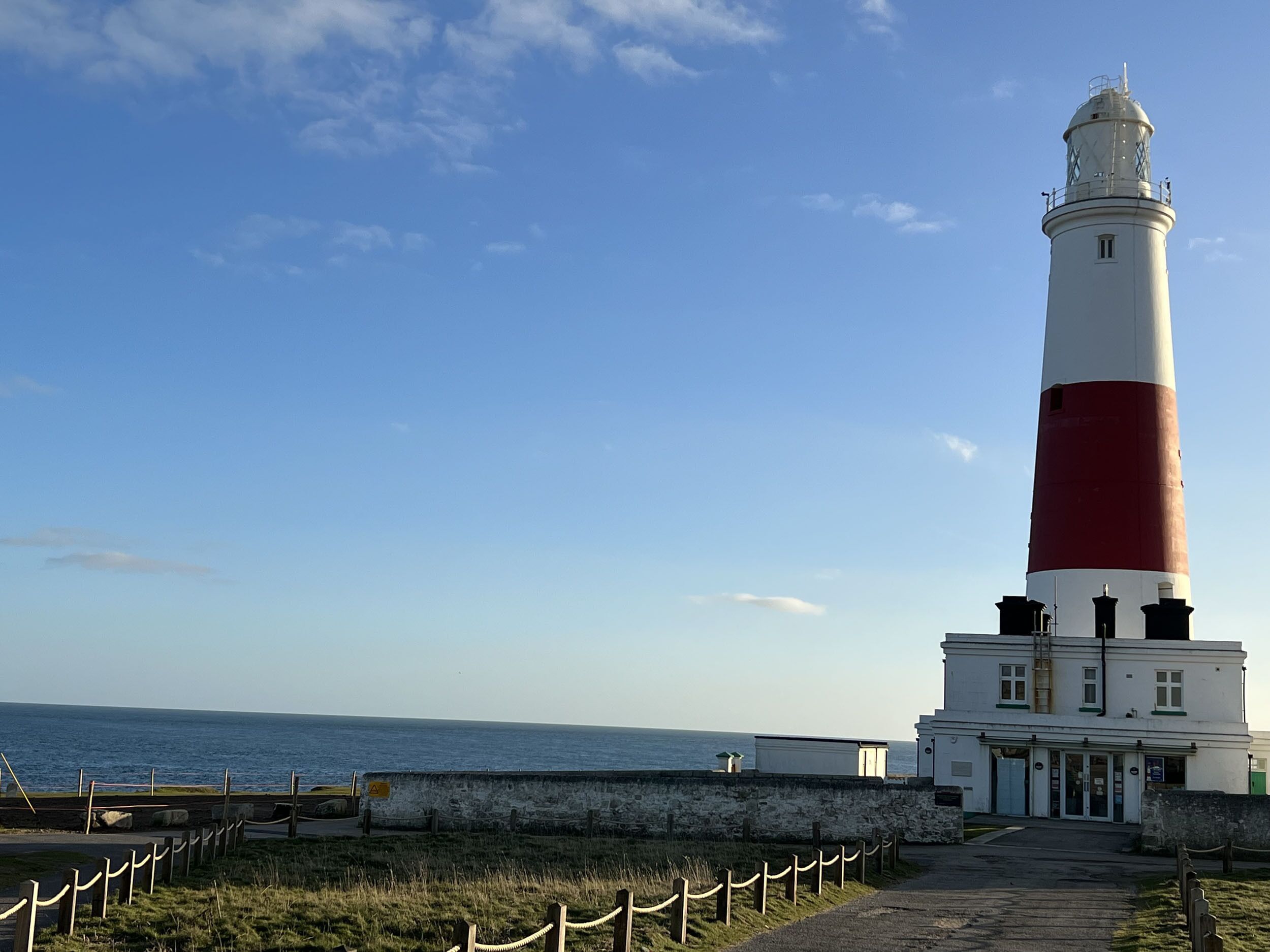 Un faro con exterior blanco y rojo, un muro de piedra y un edificio con techo verde en primer plano.