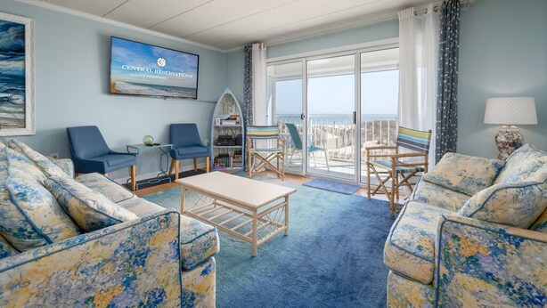 Una sala de estar con un ventanal grande, un televisor mostrando 'Central Reservations' y vista al mar.
