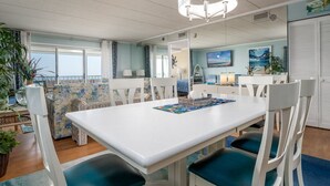 Comedor con una mesa blanca y sillas azules, una araña y vista al océano a través de amplios ventanales.