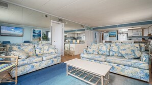 Una sala de estar con dos sofás florales, una mesa de centro y una cocina visible a través de una puerta abierta.