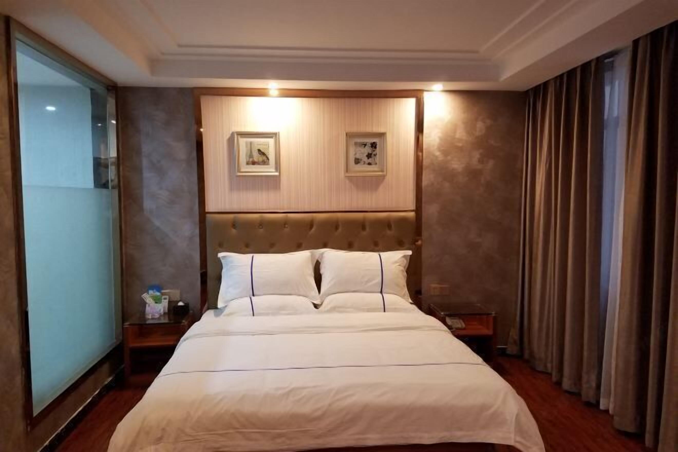 A hotel room with a large bed, a headboard with wall-mounted pictures, a bedside table, and a curtain on the window.