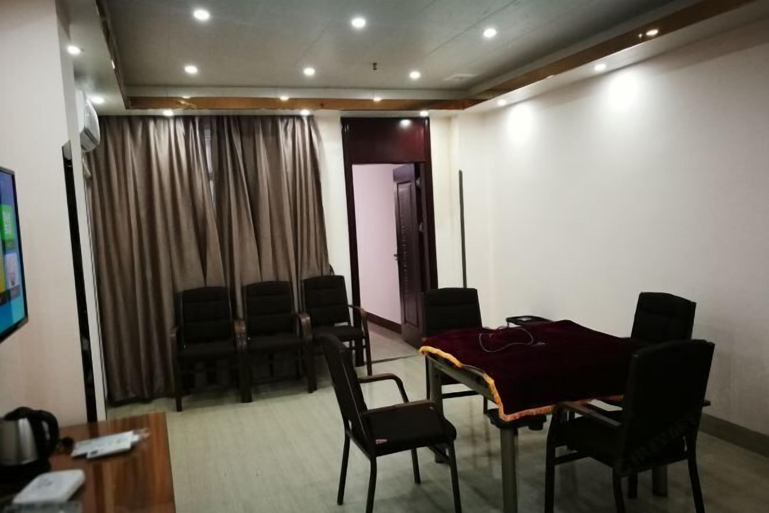 Uma sala de conferências com uma mesa de madeira, seis cadeiras e toalha de mesa vermelha. Há uma televisão à esquerda e uma porta ao fundo.