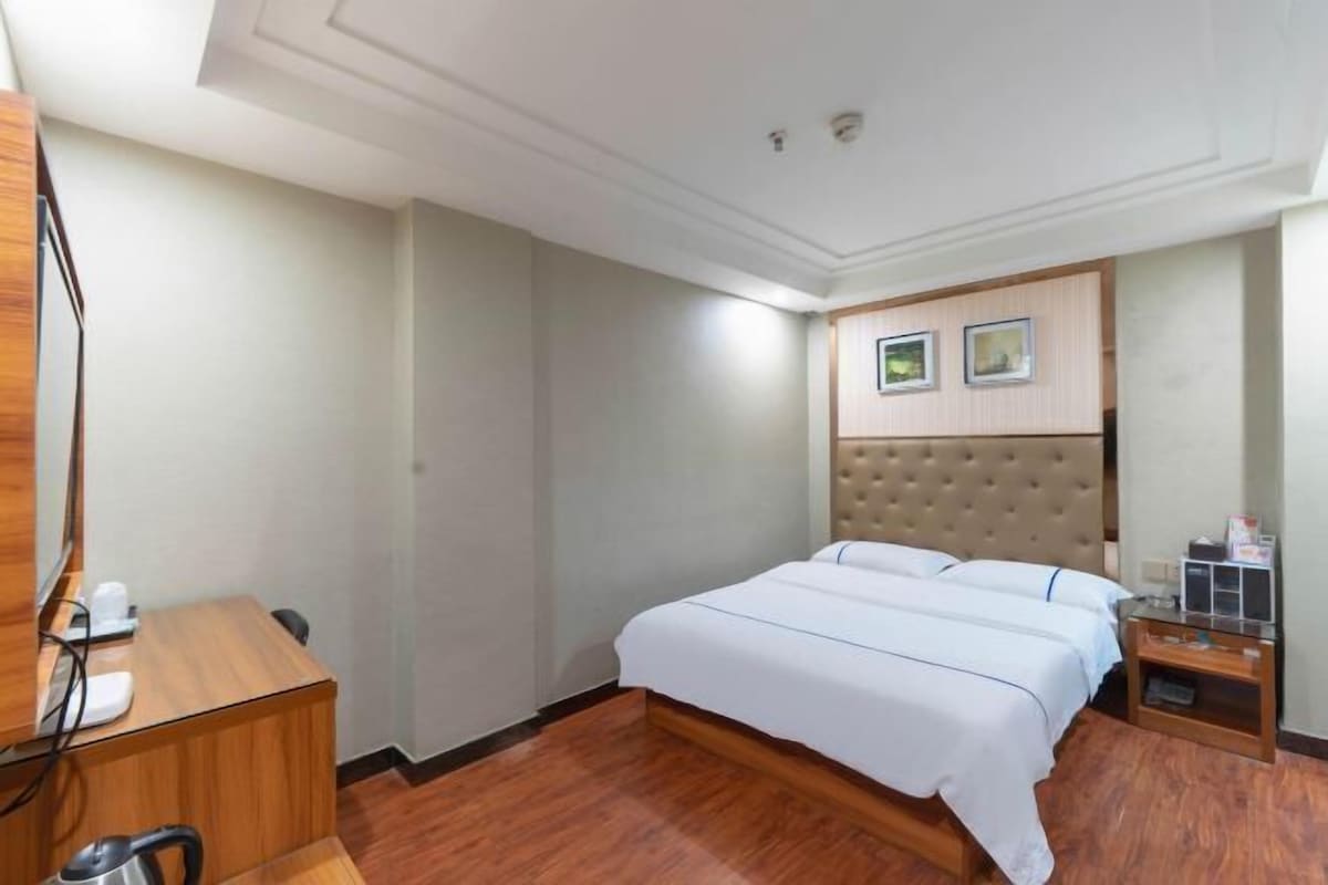 A hotel room with a bed, a desk, a chair, a coffee maker, and a small table with a lamp.