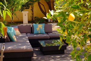 A sunlit outdoor seating area with a sofa and two side tables, surrounded by greenery and a yellow wall.