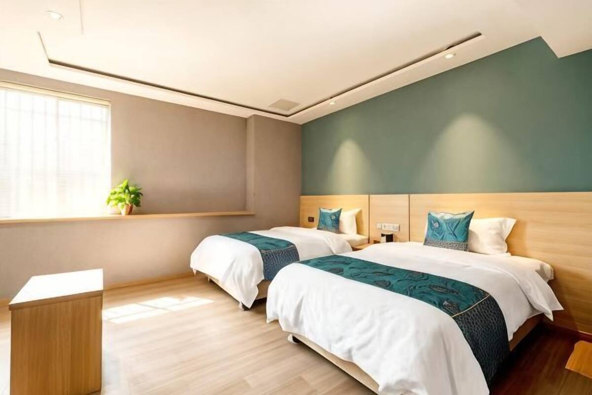 Quarto de hotel com duas camas, uma mesinha de cabeceira, janela com planta e chão de madeira.