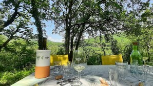 Mesa al aire libre dispuesta para comer, con copas de vino, una botella y un destornillador de corcho, rodeada de follaje y árboles.