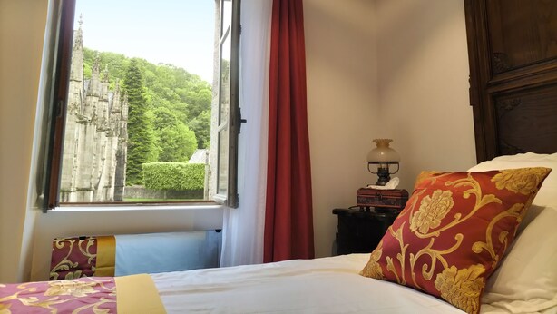 A bedroom with a large window, a bed with white and red pillows, a nightstand with a lamp, and a view of a castle through the window. - Medieval Getaway (Fougères)