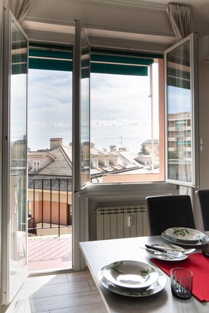A dining area with a table set for two, a view of the cityscape through large windows, and a balcony with a railing. - Sturla Modern Apartment by Wonderful Italy (Genova)