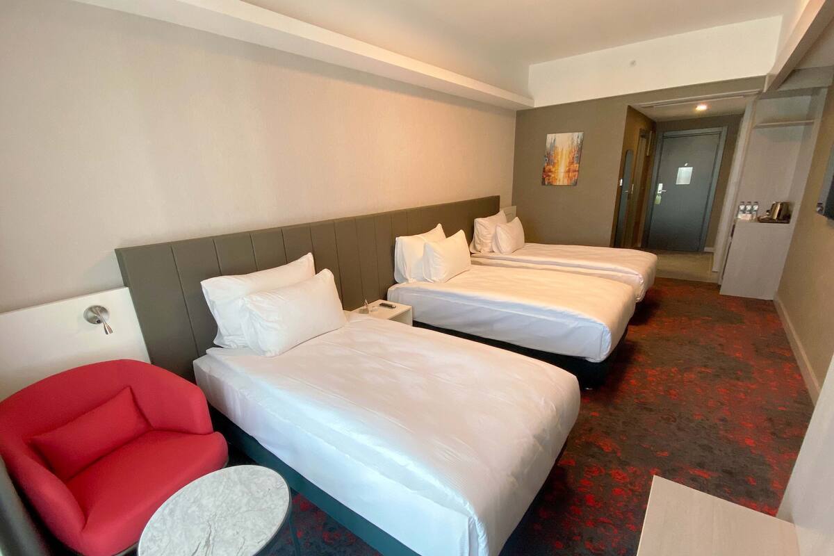 A hotel room with two beds, a red armchair, a small round table, and a door leading to a bathroom.