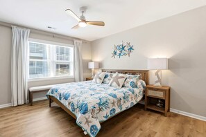 Une chambre à coucher comprenant un lit, une table de chevet, un banc, un ventilateur de plafond, une fenêtre avec des rideaux et un élément décoratif mural.
