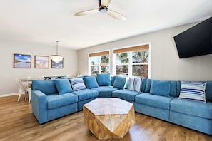 A living room with a sectional sofa, a wooden coffee table, a ceiling fan, a TV, and framed pictures on the wall. - Filling Beachy by Brunswick Vacation Rentals: Oceanfront Complex with Pool (Ocean Isle Beach)