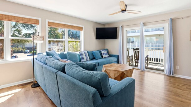 Un salon comprenant un canapé d’angle, une table basse en bois, un ventilateur de plafond, un téléviseur fixé au mur et un balcon avec une balustrade.
