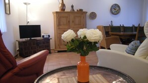 A living room with a television, a cabinet, a table with a vase of flowers, and a chair. - Bungalow 23 for up to 4 people at the RHÖN RESIDENCE vacation complex. (Dipperz)